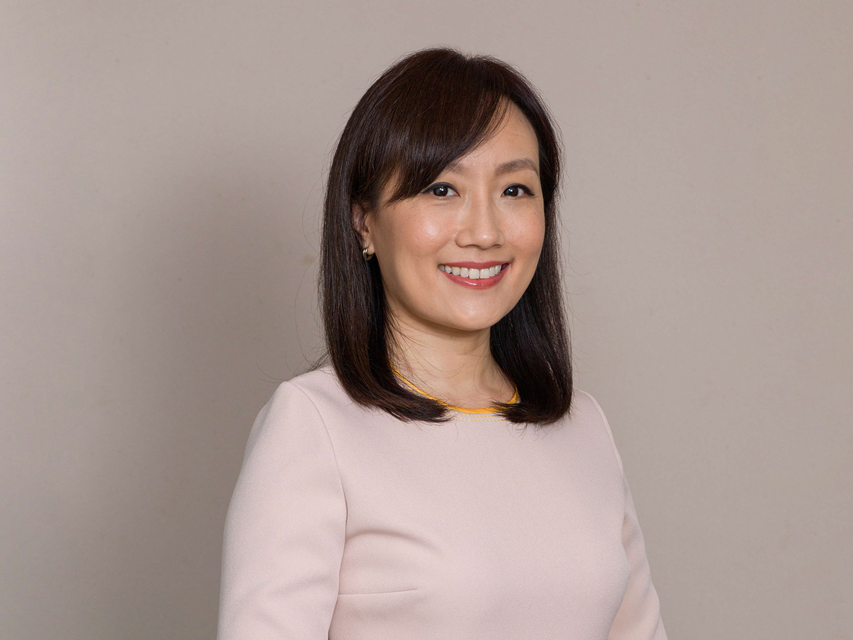 Ms Beatrice Chong in a pale pink dress smiling with hands clasped in front against a neutral beige background.